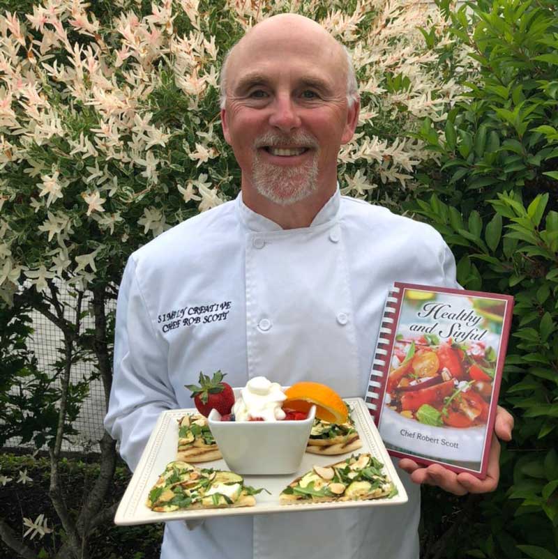 smiling man in white chef jacket holding plate of food and cookbook